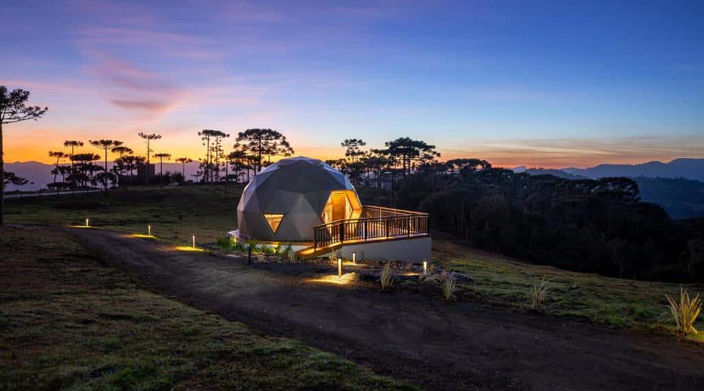 Uma cabana geodésica iluminada em uma paisagem montanhosa durante o pôr do sol. Árvores altas envolvem a estrutura de cúpula, que possui uma varanda elevada com vista para o cenário natural. Essa acomodação única permite que os hóspedes apreciem a beleza natural do local, no Orion Glamping Hospedagem Domo Geodésico, para representar onde ficar na Serra Catarinense.