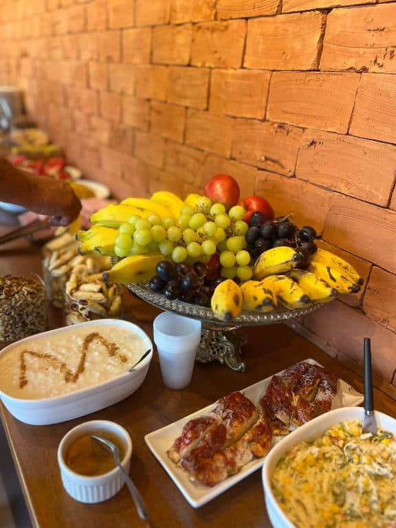 Cesta de frutas frescas e variadas, como bananas, uvas, tomates e pimentões, sobre uma mesa de madeira rústica com parede de tijolos ao fundo. é no Paraíso Capitólio Suítes Pousada. Ilustra onde ficar na serra da canastra