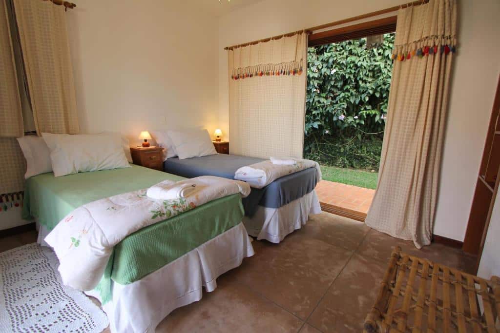 Quarto aconchegante na Pousada Asa do Vento com duas camas de solteiro, mesa de cabeceira e cortinas beges com vista para área externa com vegetação. Ilustra as pousadas em São Luiz do Paraitinga.