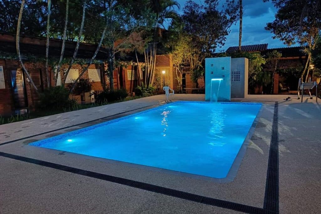 Piscina iluminada de noite na pousada barcelos, com jardim e vegetação tropical ao redor. Uma fonte d'água emite jatos de água ao lado da piscina.