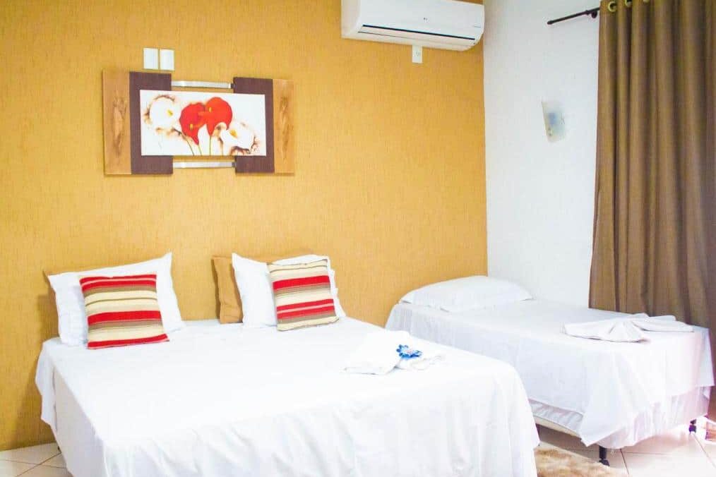 Quarto aconchegante da Pousada Encantos do Glória com ar-condicionado, decorado em tons de amarelo. Duas camas brancas com travesseiros listrados em vermelho e bege. Quadro colorido na parede exibindo flores vermelhas e amarelas.