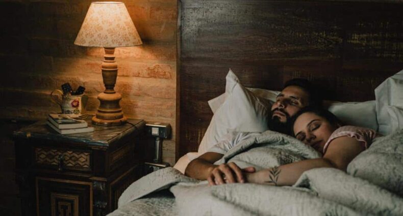 Casal dormindo abraçado na cama com roupa de cama branca e abajur aceso no quarto