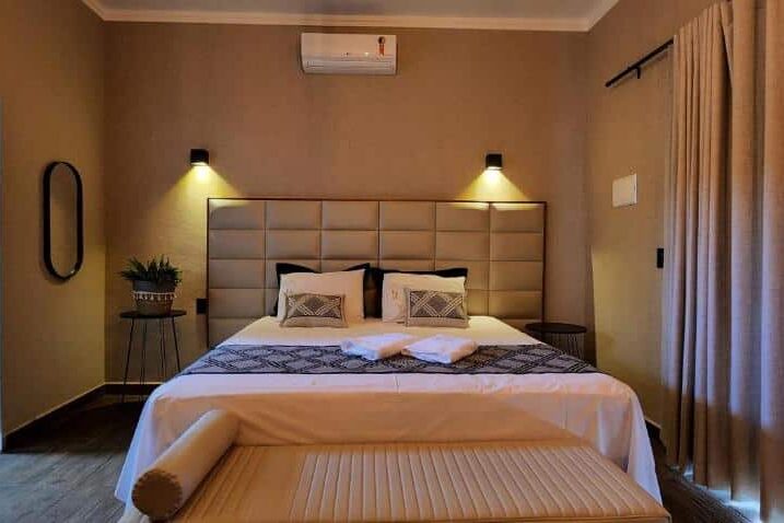 Quarto da pousada lago azul moderno com cama king size, cabeceira estofada bege, luminária de parede e janelas com cortinas.