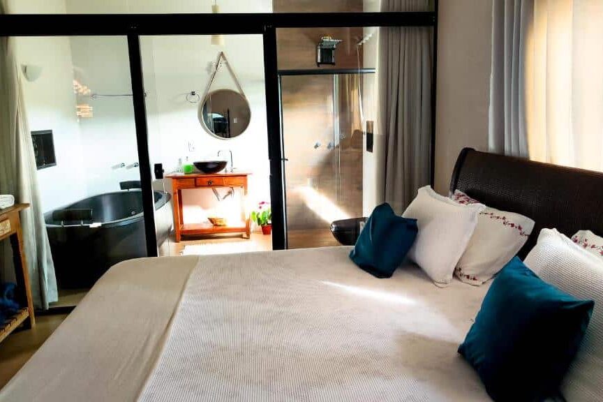 Quarto da Pousada Nascente do São Francisco com grande cama de casal, almofadas de tons escuros, espelho redondo na parede e vitrine com objetos decorativos, em ambiente aconchegante e iluminado.