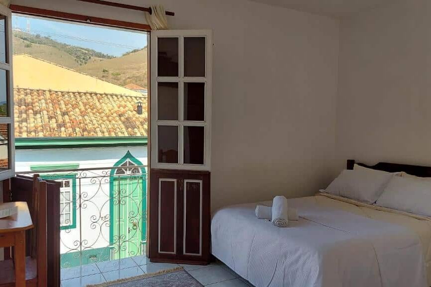 Quarto da Pousada Nativa's com vista para telhados de telhas inclinadas em uma cidade no interior, com janela aberta e mesa de madeira com cadeira