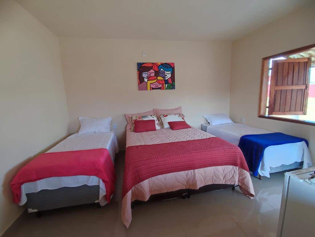 Quarto aconchegante da Pousada Pato Mergulhão com cama de casal e duas camas de solteiro, decorado com quadro colorido e almofadas vibrantes, transmitindo um ambiente agradável e convidativo.