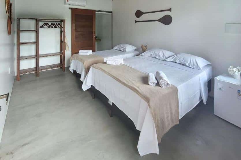 Quarto da Pousada Tauari com teto rústico, duas camas de solteiro com lençóis claros, cabideiro de madeira e luminária de teto decorativa.