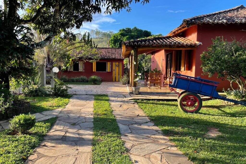 Casa de campo rústica com telhado de telhas vermelhas da Pousada Vale dos Diamantes, em meio a jardim com grama verde e vegetação exuberante. Carrinho de mão antigo azul com detalhes em vermelho está no primeiro plano, em um caminho de pedras.