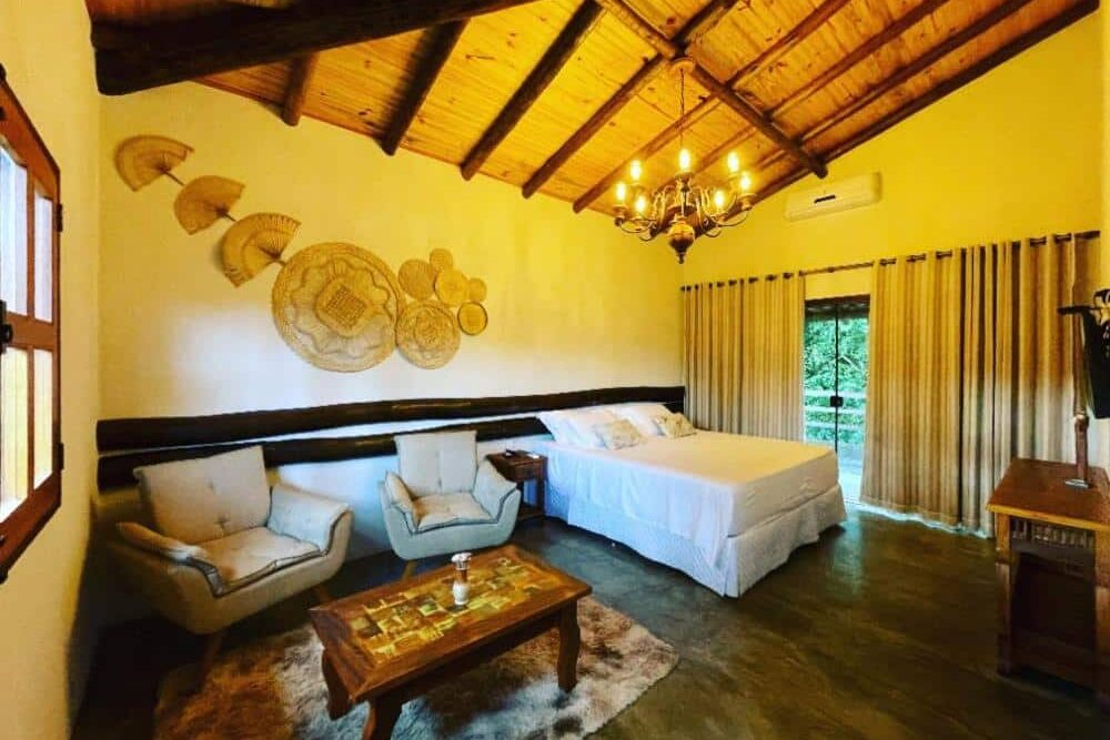 Quarto rústico da Pousada Vale dos Diamantes com teto de madeira, móveis de madeira e decoração de parede composta por objetos cestos e rendas, criando um ambiente aconchegante e acolhedor.