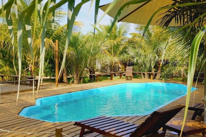Um lugar verde na Pousada Vila Araticum com uma piscina azul cercada por uma área de descanso com cadeiras de madeira e palmeiras abundantes. Ilustra onde ficar em Santo André na Bahia.