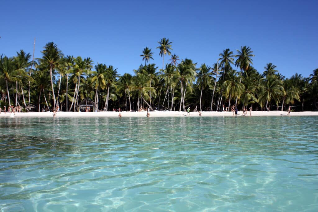 Uma das praias de Isla Saona com mar azul, pessoas andando na areia branca e muitas palmeiras cercando a praia.