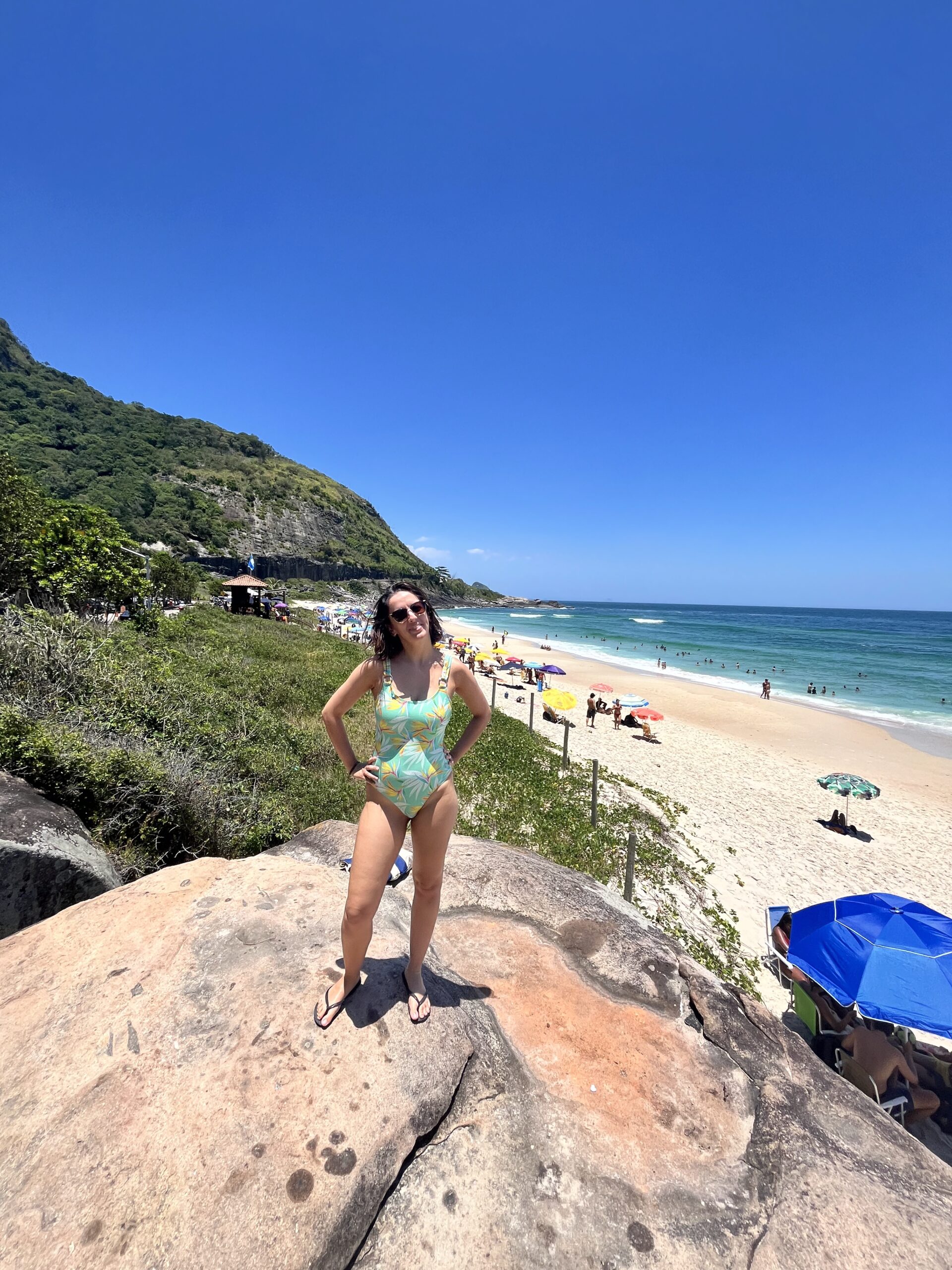 Mulher em maiô brilhante curtindo praia de Itacoatiara, Niterói, com paisagem de montanhas, céu azul e mar turquesa ao fundo.