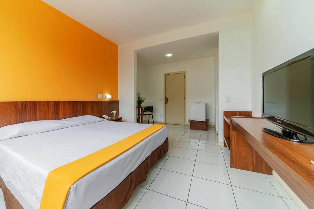 Quarto do Alice Vitoria Hotel Arte by Nobile com parede laranja, cama grande, móveis de madeira e televisão na parede.