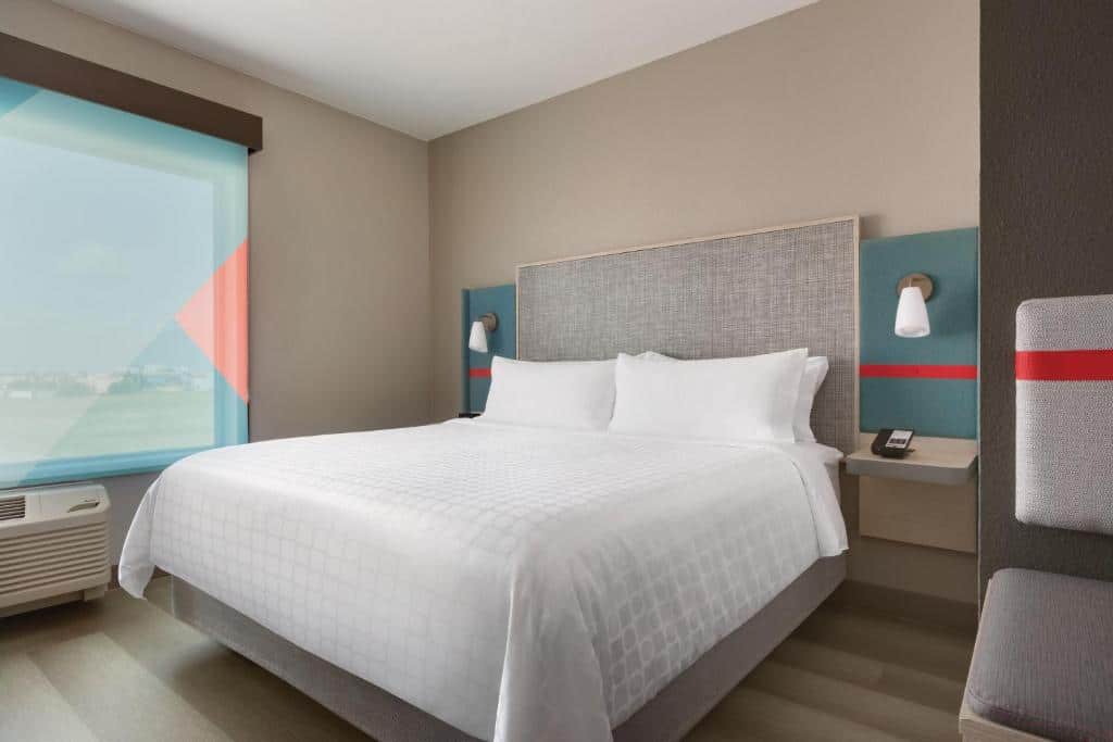 Quarto elegante do avid hotel com paredes neutras, cama de casal com cabeceira ripada e detalhes em vermelho e azul, mesitas de cabeceira e ar-condicionado.