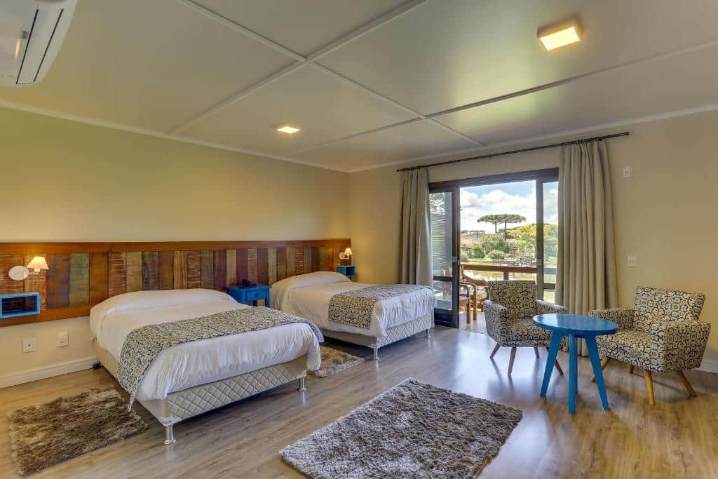 Quarto aconchegante com duas camas de casal, parede de madeira rústica, cortinas e mobília estampada, além de mesa e cadeiras em um canto da sala com vista para área externa, no Boqueirão Hotel Fazenda, para representar onde ficar na Serra Catararinense.