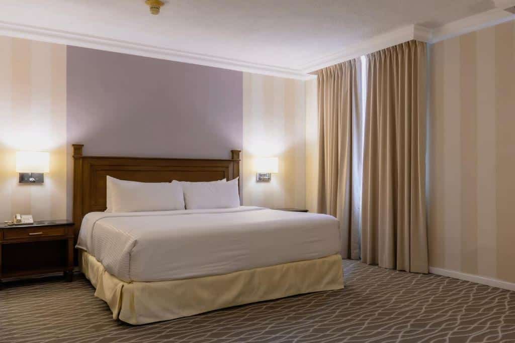 Quarto do Gamma Monterrey Gran Hotel Ancira com cama de casal, cabeceira de madeira escura e cortinas bege. Ambiente iluminado e espaçoso, com piso de carpete com design orgânico.