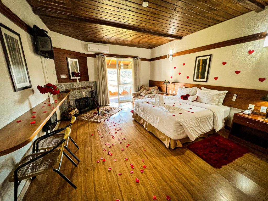 Quarto romântico com flores de pétalas vermelhas espalhadas pelo piso de madeira, lareira de pedra e grandes janelas revelando vista externa. Cama de casal com lençóis claros e decoração luxuosa em tons de vermelho e marrom, no Hotel Fazenda Rota dos Cânions, para representar onde ficar na Serra Catarinense.