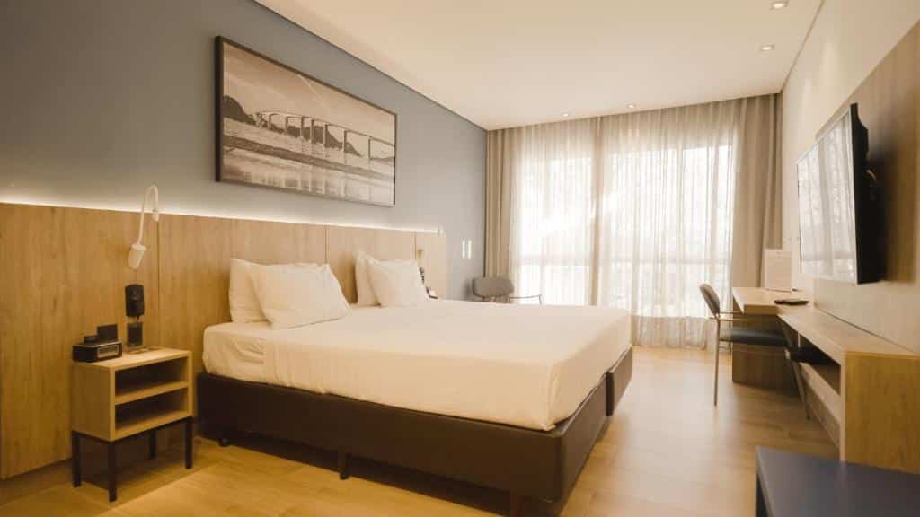 Quarto do Hotel Senac Ilha do Boi com móveis de madeira, cama de casal branca e parede cinza com quadro paisagístico.