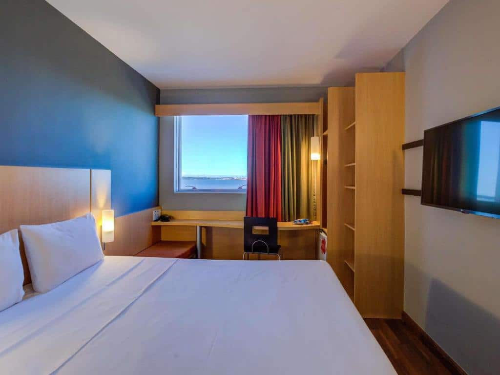 Quarto do ibis Vitoria perto da Praia de Camburi com vista panorâmica da praia e do mar, móveis de madeira clara, cortinas vermelhas e TV de tela plana.
