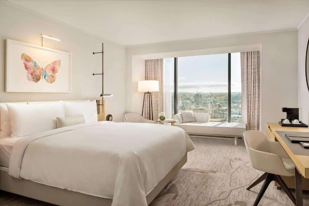 Quarto do JW Marriott Parq Vancouver com vista panorâmica da cidade, contendo cama king size, poltrona, escrivaninha e obra de arte com borboleta colorida na parede.