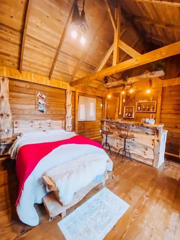 Cabana de madeira acolhedora com iluminação aconchegante, cama com cobertor vermelho e roupas de cama claras, balcão rústico com objetos decorativos, na Pousada Flocos de Neve, para representar onde ficar em Urubici.