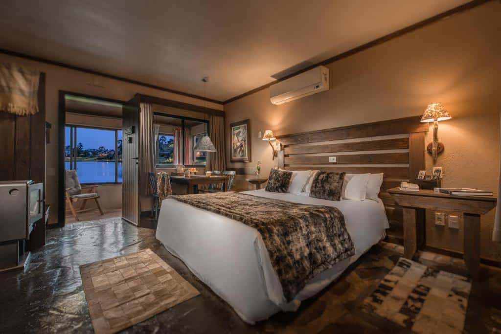 Quarto rústico e aconchegante no Rio do Rastro Eco Resort com vista para o lago, cama grande, decoração em madeira e tecidos quentes, luminárias de parede e detalhes em tons terrosos, para representar onde ficar na Serra Catarinense.