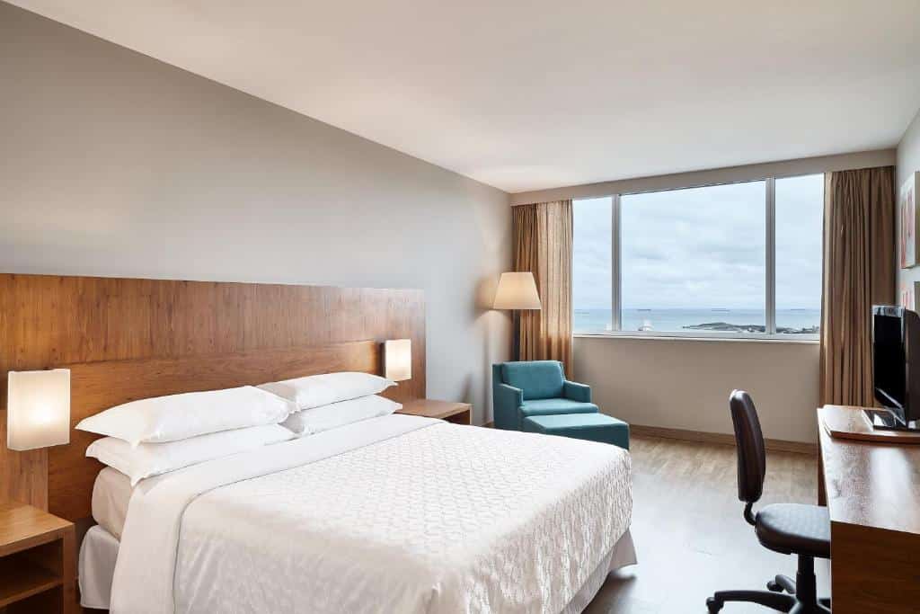 Quarto do Sheraton Vitoria com ampla janela panorâmica e vista para o mar. Cama king size com roupa de cama branca, poltrona azul turquesa, escrivaninha de madeira e televisor na parede.