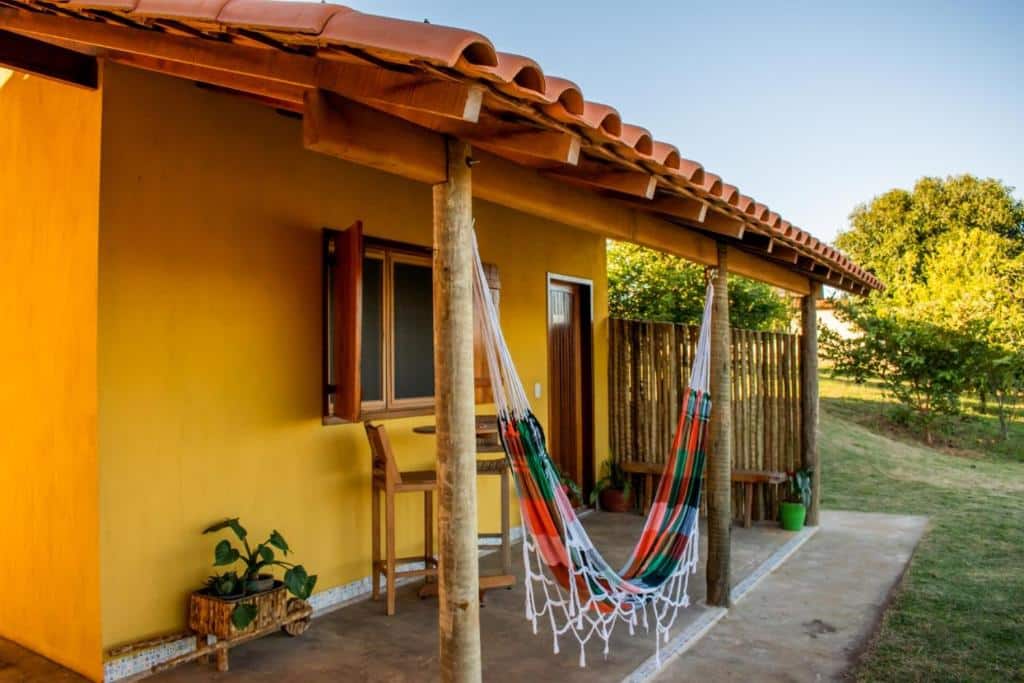 Casa rústica amarela com telhado de telhas terracota e varanda com rede de descanso colorida, cercada por vegetação verde em ambiente bucólico. é o Refúgio Pé da Serra – Chalés