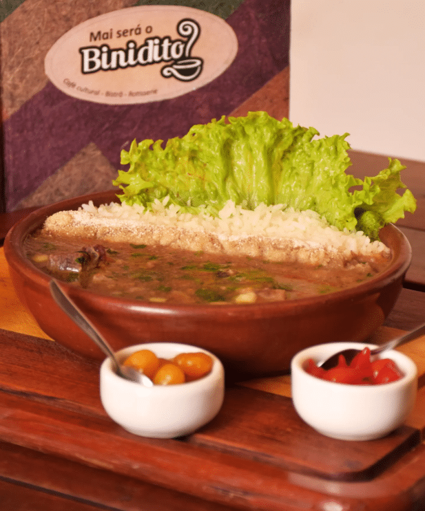 Prato de sopa de feijão em tigela de cerâmica com folhas de alface fresca, acompanhado de pequenos bowls contendo fatias de laranja e pimentos vermelhos, em uma superfície de madeira e logotipo do "Binidito" ao fundo.