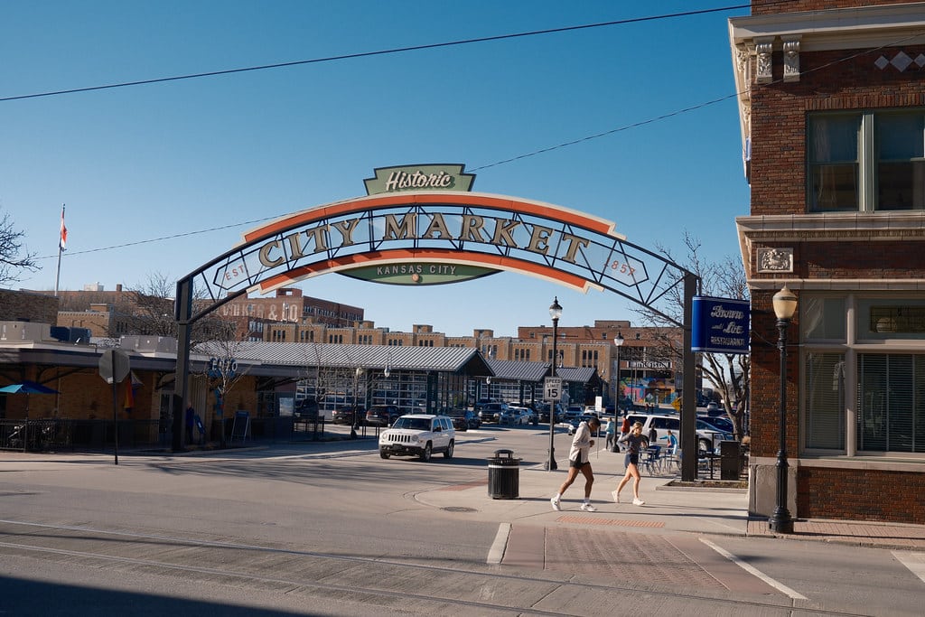 Arco de entrada do Mercado Público Histórico de Kansas City, estrutura arquitetônica de tijolos vermelhos com detalhes em laranja e letreiro em estilo art déco, com pessoas caminhando pelo local em dia ensolarado.