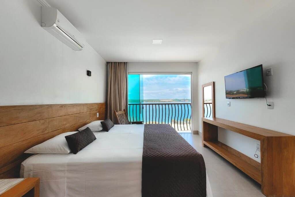 Quarto de hotel Riviera Capitólio Hotel com ampla janela com vista para praia tropical e céu azul. Cama de casal com lençóis claros e decoração minimalista em tons de madeira. Televisão de tela grande na parede e ar-condicionado. Ilustra onde ficar na serra da canastra