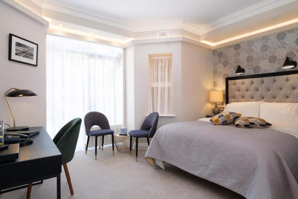 Quarto no Roxford Lodge Hotel com cama king size, cabeceira estofada cinza, duas poltronas azuis e escrivaninha preta moderna. Iluminação ambiente e paisagem externa visível por janelas amplas.