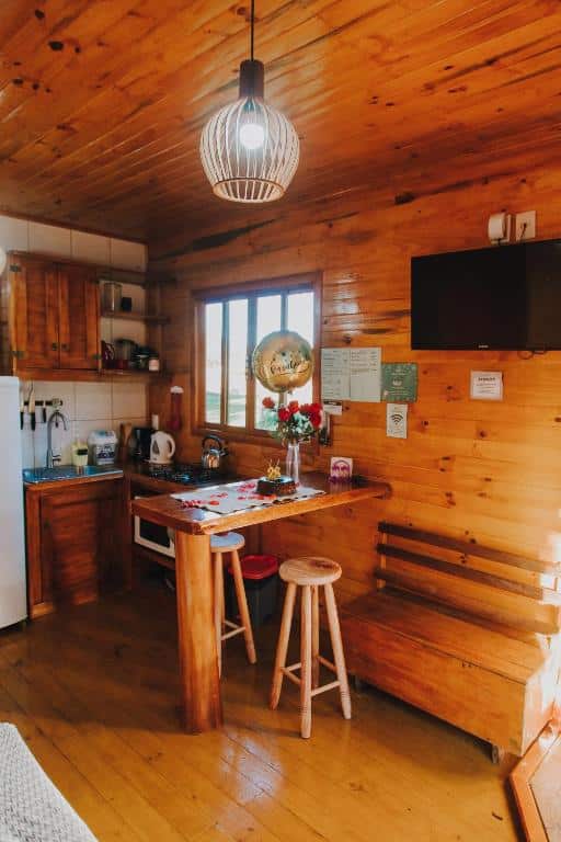 Cozinha rústica em madeira com bancada de madeira, fogão e pia. Luminária de design moderno pende do teto. Flores vermelhas decoram a cozinha aconchegante, na Casa de Campo Santa Paulina.