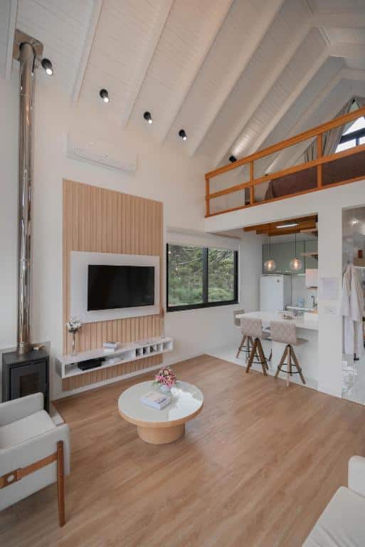 Amplo espaço interno com pé-direito alto, paredes claras e móveis de madeira em estilo rústico moderno. Uma TV de tela grande está montada na parede, com prateleiras adjacentes. Uma escada de madeira leva a um mezanino, com uma pequena cozinha aberta no fundo.