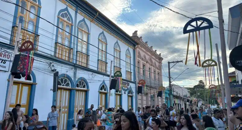 Rua movimentada com prédios históricos coloridos, bandeiras e símbolos decorativos, grupos de pessoas presentes em São Luiz do Paraitinga.