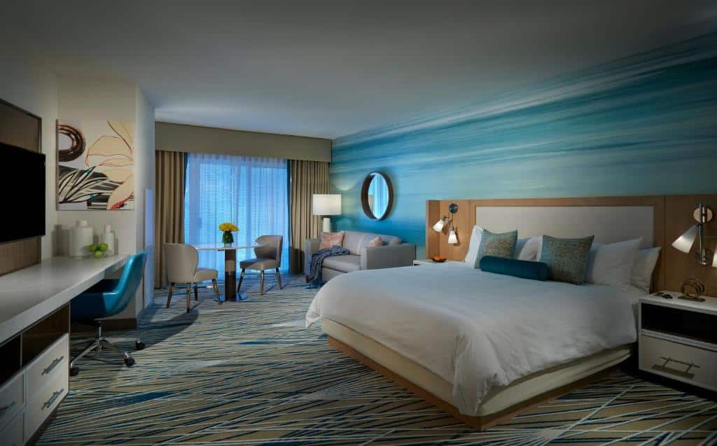 Quarto do Seminole Hard Rock Hotel & Casino Hollywood com cama king size, sofá cinza, cadeira azul-turquesa e mesa de trabalho. Ampla janela com vista para exterior com tonalidades azuladas. Essa é uma das dicas de onde ficar perto do Hard Rock Stadium.