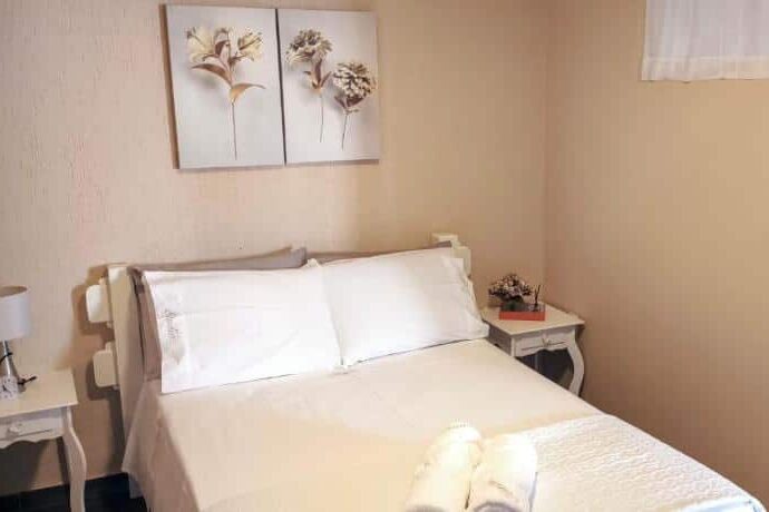 Quarto aconchegante no Studio do Wander com decoração minimalista, incluindo quadros florais na parede, cama com lençóis brancos e mesinha de cabeceira ao lado.