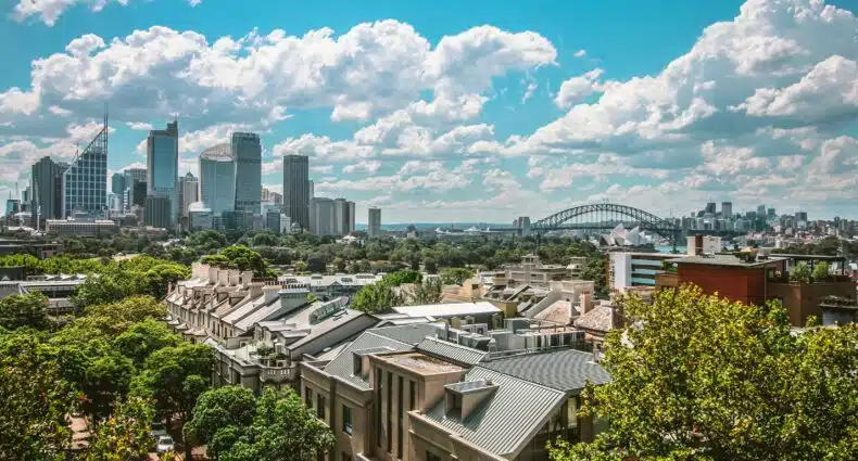 Skyline da cidade de Sydney, Austrália, com imponentes arranha-céus, a emblemática Ponte da Baía e a famosa Ópera de Sydney ao fundo, sob um céu azul pontilhado de nuvens brancas. Foto usada como capa para o post de onde ficar em Sydney.