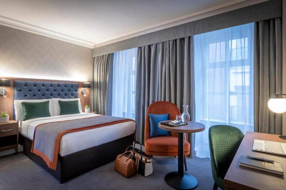 Quarto do The Fleet com cama de casal de estofado capitonê azul, mesa de canto com luminária e vaso de planta, cadeira redonda laranja e mesa de trabalho com notebook.