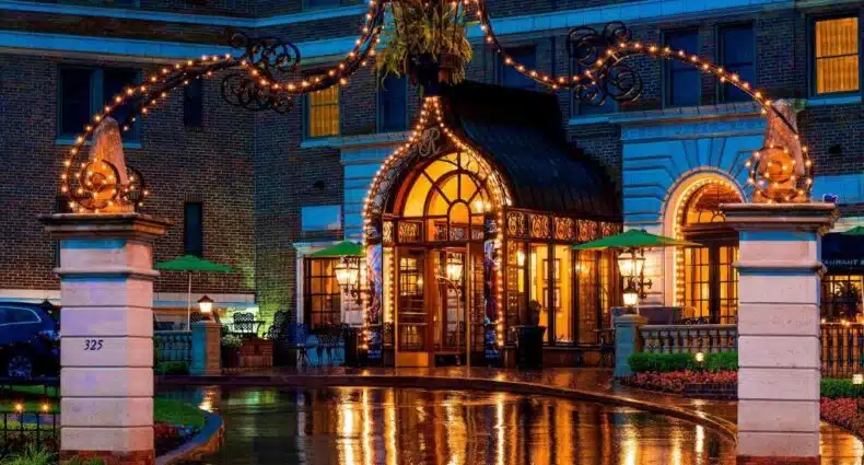 Belo edifício iluminado à noite com varandas, arcos e detalhes decorativos refletidos em poça d'água no chão em calçada ornamentada. Esse é o The Raphael Hotel, uma das indicações de onde ficar em Kansas City.