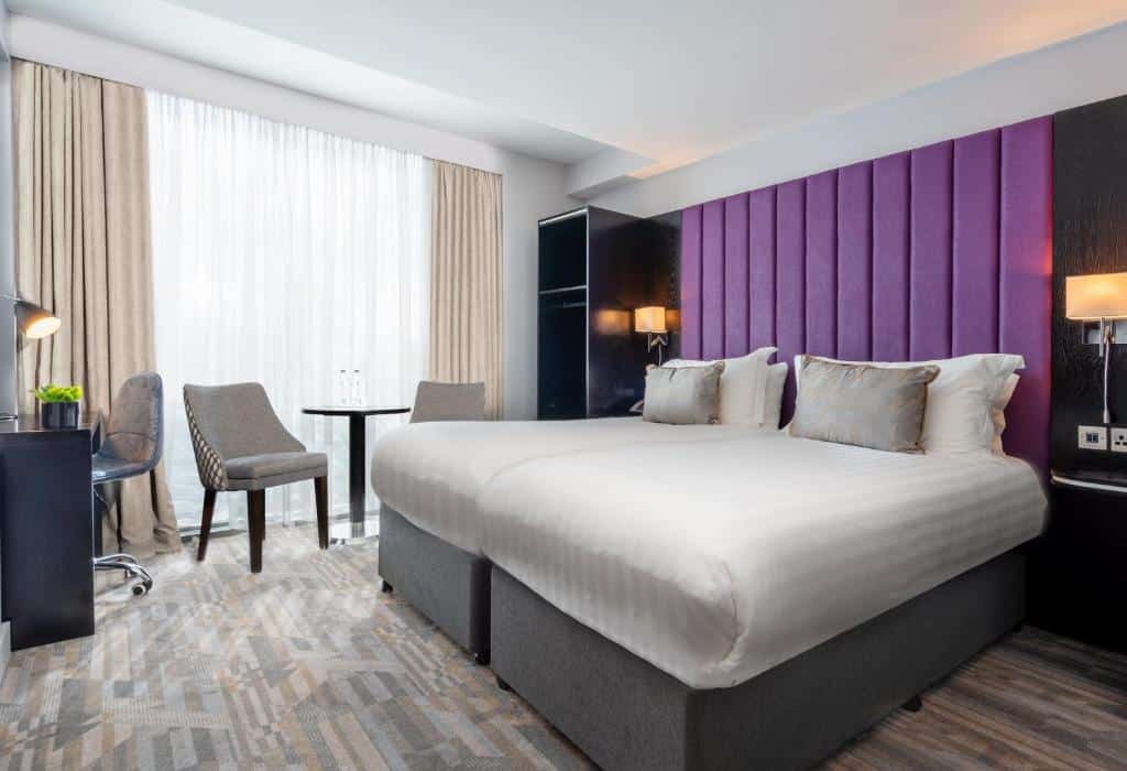 Quarto do The Trinity City Hotel com parede de cabeceira violeta, cama de casal branca, cadeira cinza e móveis escuros.