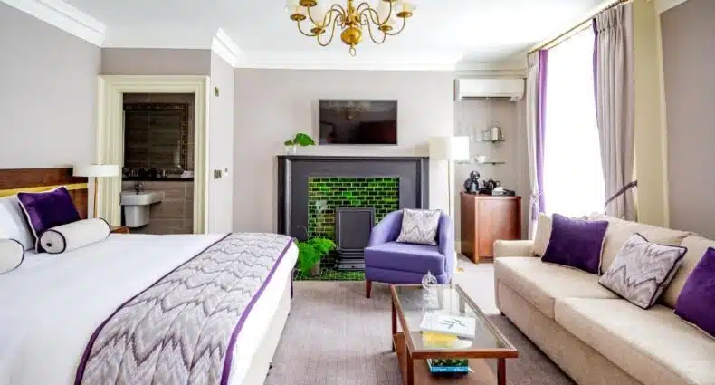 Quarto espaçoso do Trinity Townhouse Hotel com decoração elegante em tons de roxo e cinza, com cama grande, poltronas de veludo, lareira de tijolos verdes e lustre dourado no teto. A foto faz parte do post de onde ficar em Dublin.