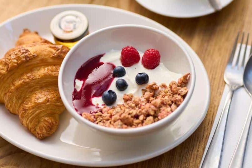 Refeição saudável e colorida no Trinity Townhouse Hotel com croissante, iogurte com frutas vermelhas, cereais granola e raspas de limão em prato branco sobre mesa de madeira.