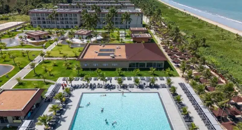 Um complexo de resort à beira-mar com vários edifícios de apartamentos, rodeado por vegetação tropical e com uma piscina central onde diversas pessoas estão nadando. Essa é a Vila Galé Resort Alagoas - All Inclusive, uma das indicações de onde ficar em Barra de Santo Antônio.