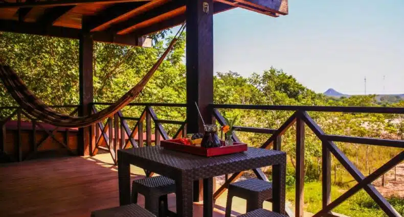 Na Villa Arumã Pousada, uma varanda rústica com rede de descanso, mesa e bancos de vime em cenário natural com árvores verdes e montanhas ao fundo. A foto é capa do post de onde ficar em Alter do Chão.