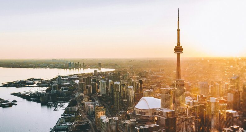 Vista aérea da skyline moderna e vibrante da cidade de Toronto, no Canadá. Destaca-se a imponente Torre CN, arranha-céu mais alto da cidade, com 553 metros de altura. A região é repleta de edifícios comerciais e residenciais. Ao fundo, é possível avistar o lago Ontário e o horizonte na luz dourada do pôr do sol.