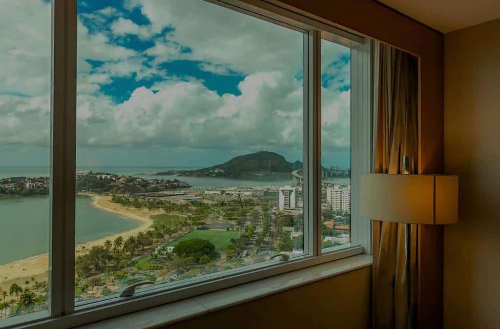 Vista panorâmica da praia com morros e edifícios ao fundo em dia ensolarado com nuvens brancas no céu azul, do Sheraton Vitoria.
