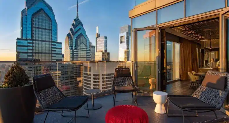 Prédios modernos de vidro e aço em centro de cidade, com área de lazer aconchegante em varanda com cadeiras e pufe vermelho, vista do terraço do W Philadelphia, para representar onde ficar na Filadélfia.