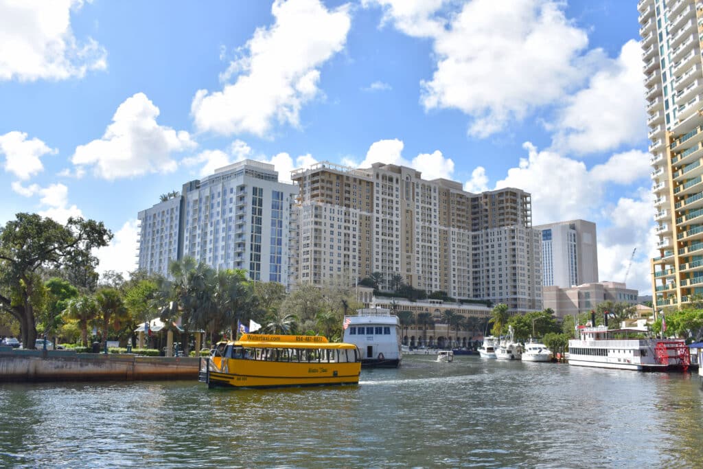 Prédios e hotéis de luxo à beira de rio em centro urbano movimentado com barco de turismo amarelo navegando nas águas. Imagem para ilustrar post sobre o que fazer em Fort Lauderdale.