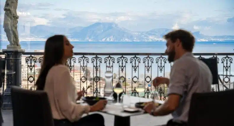 Casal apreciando a vista da bela paisagem litorânea de Nápoles com montanhas ao fundo durante refeição em varanda com balaustrada ornamentada, no Grand Hotel Parker's. Representa onde ficar em Nápoles.